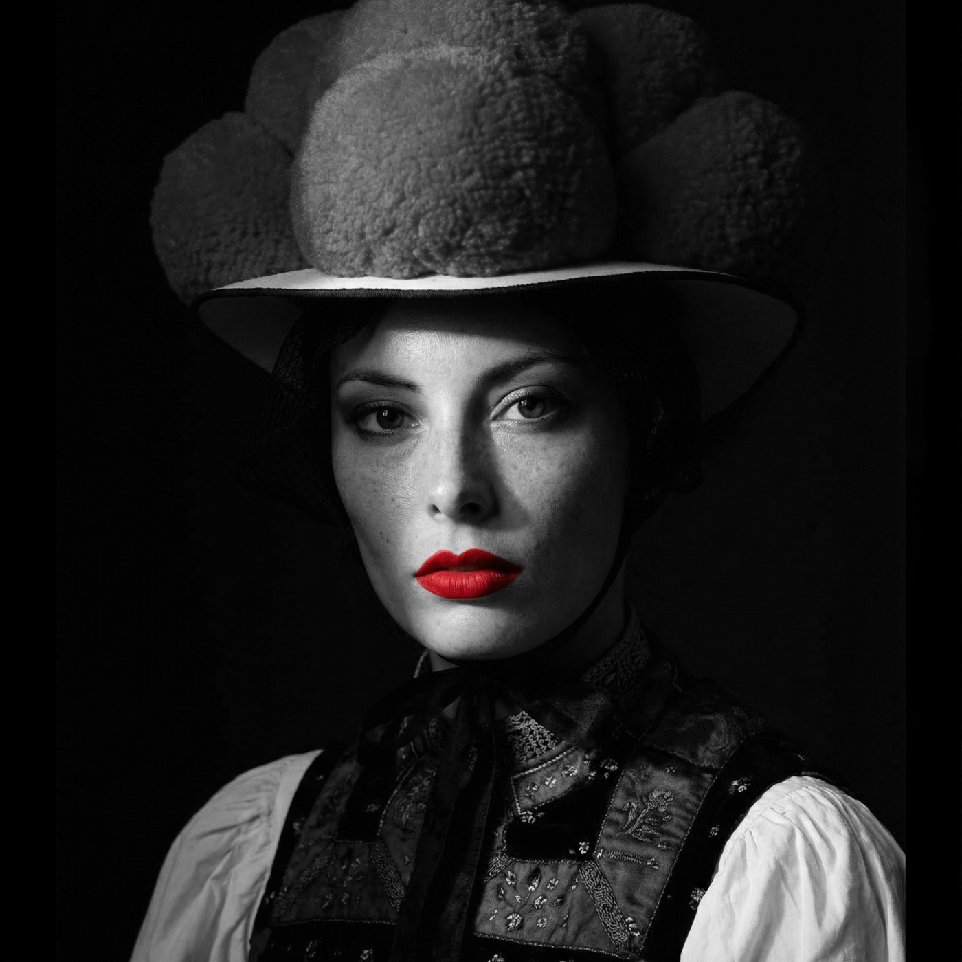 Porträt einer Frau mit Schwarzwälder Bollenhut in Schwarz-Weiß mit intensiv rot hervorgehobenen Lippen vor dunklem Grund.