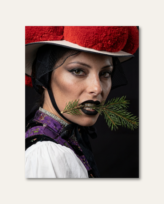 Nahaufnahme einer Frau in Schwarzwälder Tracht mit rotem Bollenhut und dunklem Lippenstift, die auf einem grünen Tannenzweig beißt. Das ausdrucksstarke Porträt besticht durch den dramatischen Kontrast zwischen dunklem Hintergrund, leuchtend rotem Bollenhut und frischem Tannengrün.