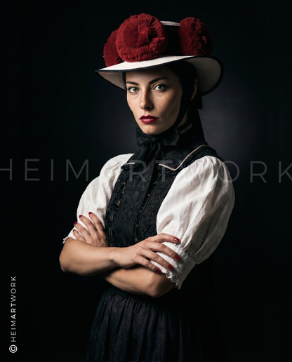 Das Motiv zeigt ein ausdrucksstarkes Porträt einer Frau in traditioneller Schwarzwald-Tracht mit dem ikonischen Rosenhut vor einem dunklen, atmosphärischen Hintergrund. Die Kombination aus historischem Gewand und moderner, fast schon trotziger Pose verleiht dem Bild eine kraftvolle, zeitgemäße Ästhetik.